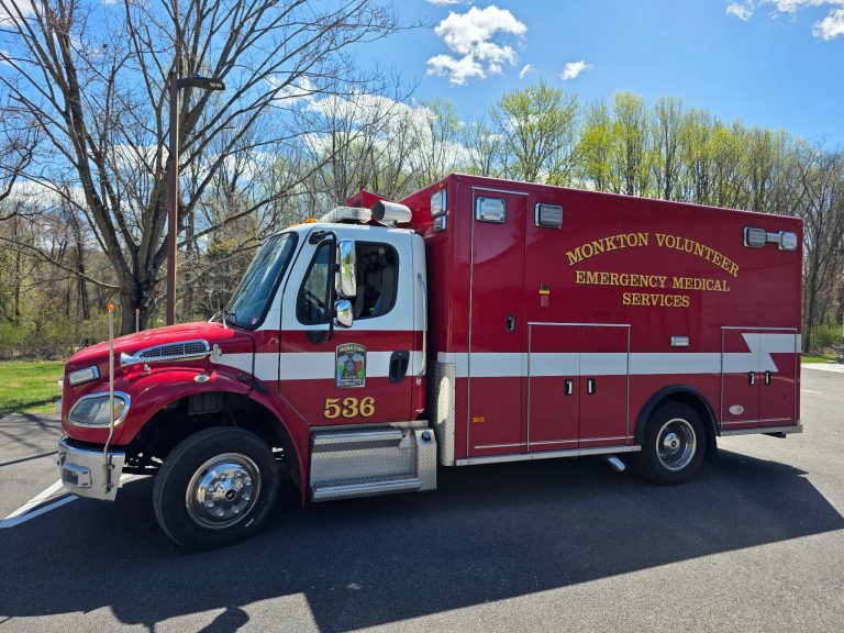 A red Monokton Volunteer Emergency Medical Services ambulance with the number 536 is parked on pavement near leafless trees and green foliage under a blue sky with clouds.
