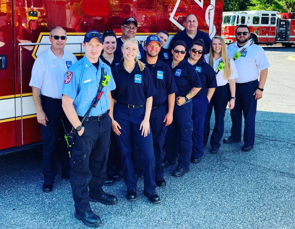 A group of emergency responders, including both men and women, stand in front of a fire truck. They wear uniforms with patches and badges. Some smile at the camera, and one person wears a blue uniform with a body camera. Sunlight illuminates the scene.