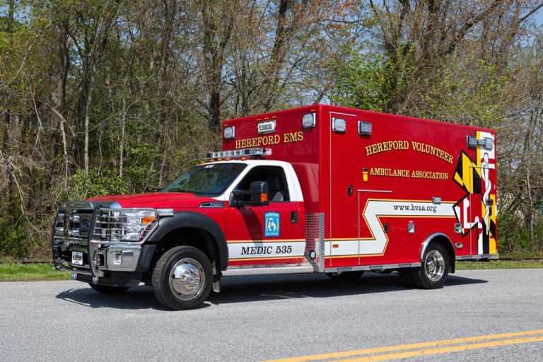 A red Hereford Volunteer Ambulance Association emergency vehicle is parked on a road beside trees and greenery, with "Hereford EMS" and "Medic 535" written on the side.