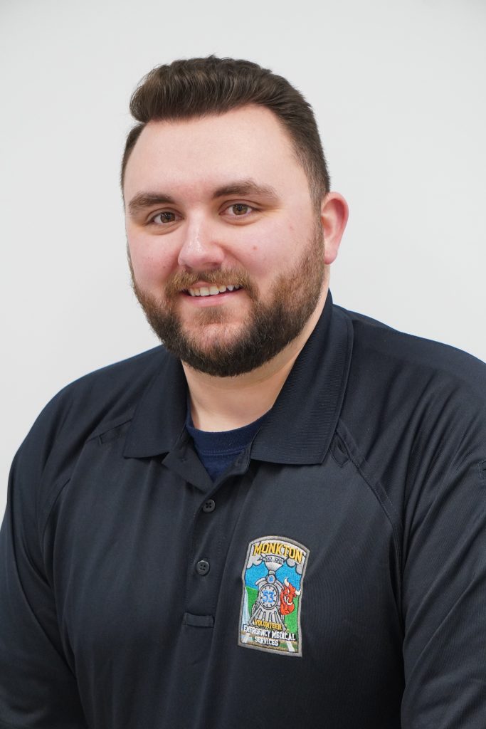 A man with short brown hair and a beard smiles at the camera. He is wearing a dark blue polo shirt with a Moncton Fire and Emergency Services logo on the left chest. The background is plain and white.