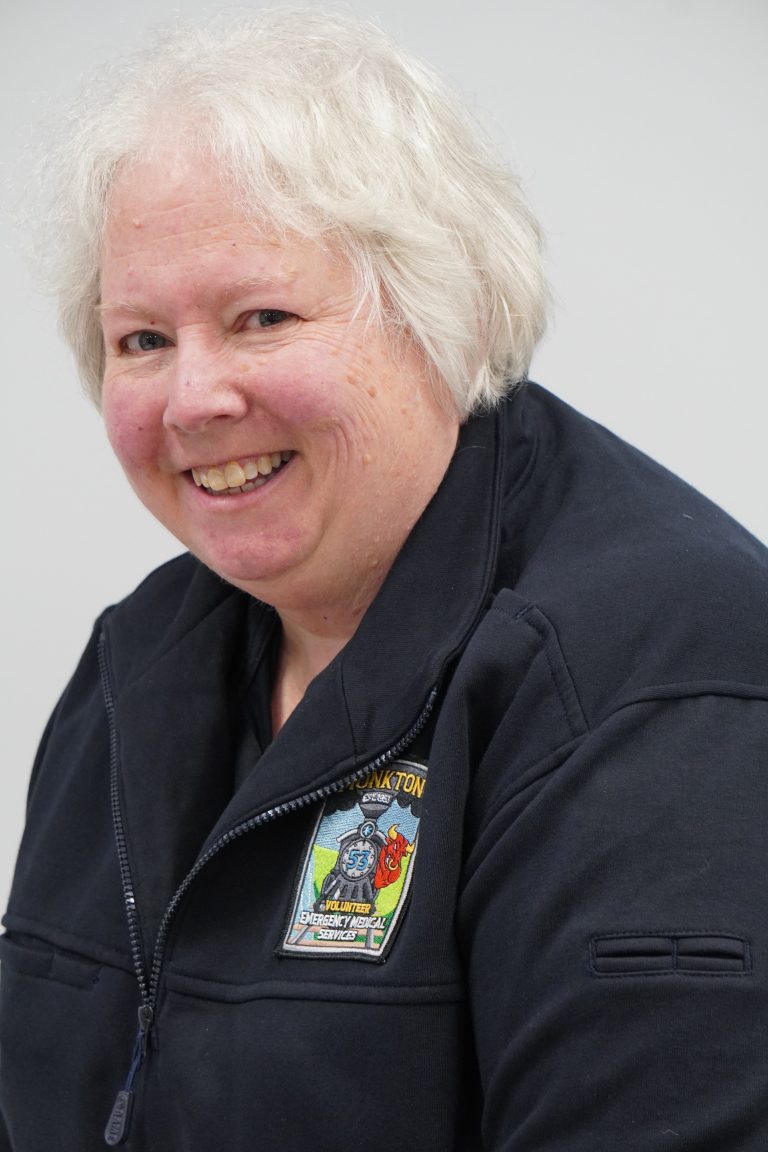 A person with short white hair smiles at the camera. They are wearing a dark jacket with a colorful embroidered patch on the chest. The background is plain and light-colored.