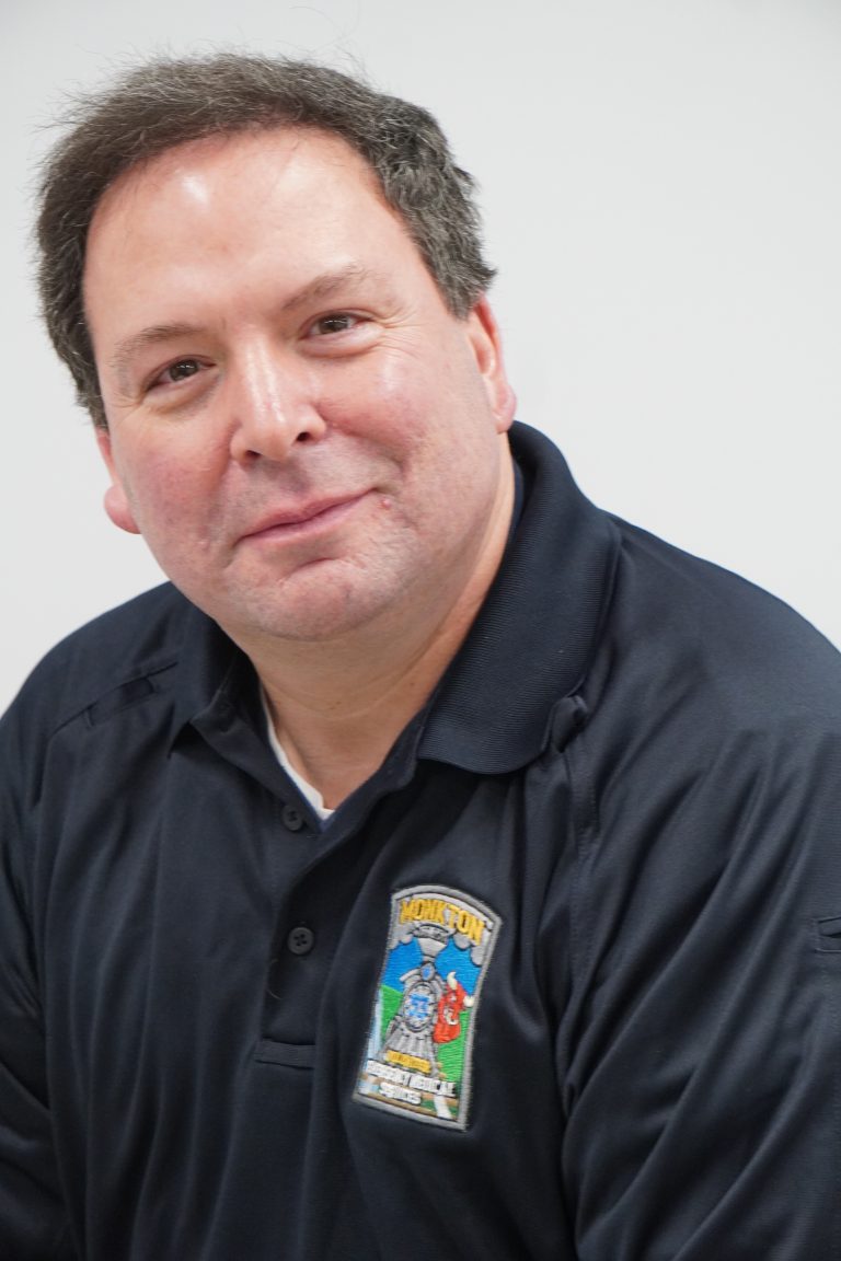 A man with short dark hair, wearing a navy blue collared shirt with a colorful embroidered patch, smiles gently at the camera against a plain white background.