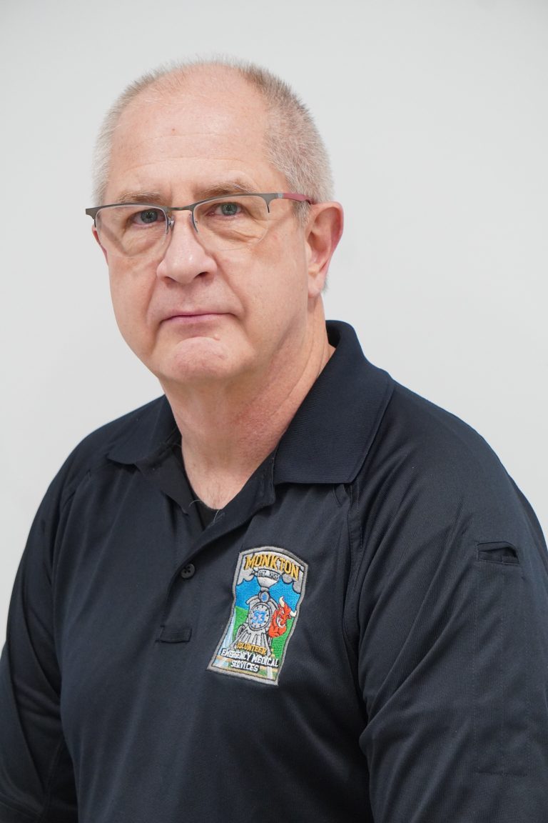 A middle-aged man with short gray hair and glasses wears a black collared shirt with a colorful patch that says "Hometown" on the chest. He is looking at the camera against a plain white background.