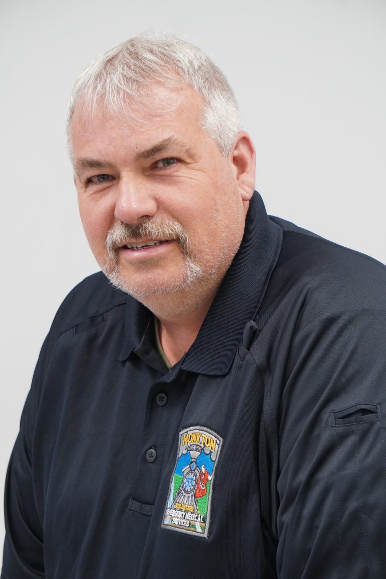 A middle-aged man with short gray hair and a mustache is wearing a dark navy shirt with a colorful embroidered patch on the chest. He is smiling slightly and posed against a plain light background.