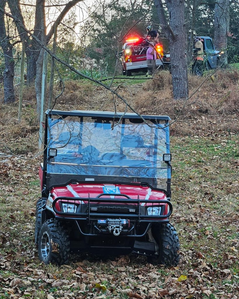 A red utility vehicle is parked on a leaf-covered slope in the foreground, while a fire truck with flashing lights and several firefighters is visible in the background among trees.