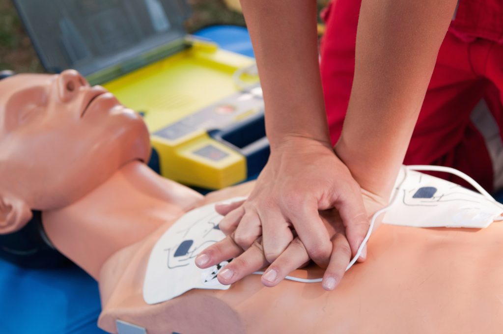 A person practices CPR chest compressions on a medical training mannequin, with defibrillator pads attached and an automated external defibrillator (AED) visible in the background.