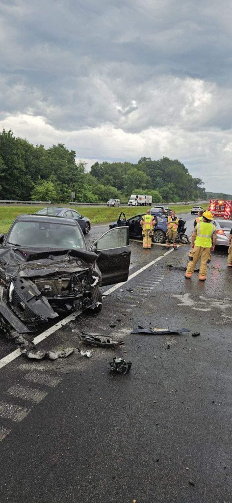 Two badly damaged black cars after a crash on a wet highway. Emergency responders in yellow gear stand nearby. Debris is scattered across the road, and emergency vehicles are in the background under cloudy skies.