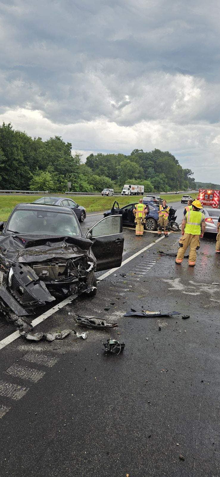 Two badly damaged black cars after a crash on a wet highway. Emergency responders in yellow gear stand nearby. Debris is scattered across the road, and emergency vehicles are in the background under cloudy skies.