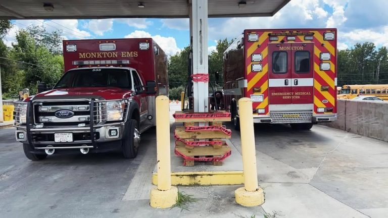 Two Monkton EMS emergency vehicles are parked under a canopy at a service station. A yellow pole and a metal structure wrapped with red caution tape stand between them. Trees and school buses are visible in the background.