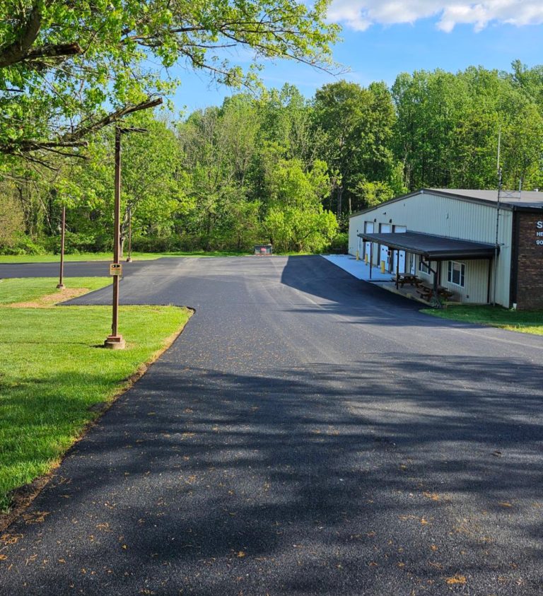 A freshly paved asphalt parking lot beside a metal building, surrounded by green trees and grass under a partly cloudy blue sky. A lamp post stands along the edge of the parking area.