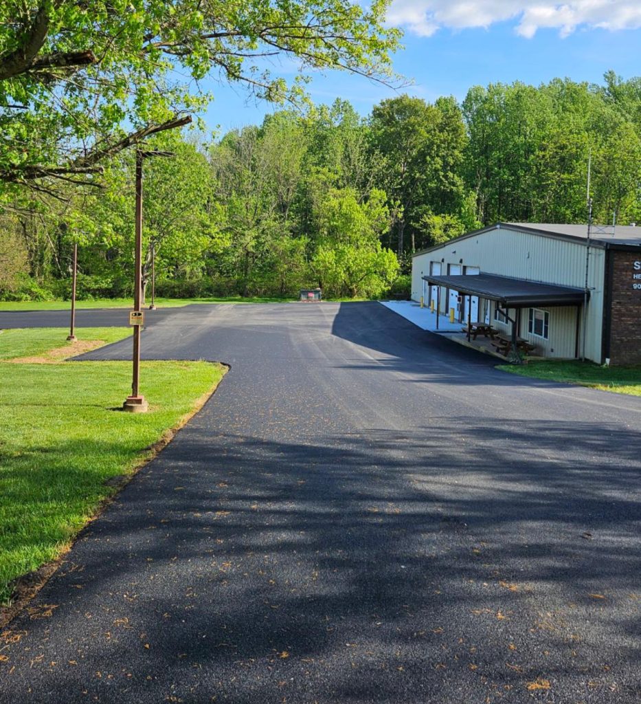 A freshly paved asphalt parking lot beside a metal building, surrounded by green trees and grass under a partly cloudy blue sky. A lamp post stands along the edge of the parking area.