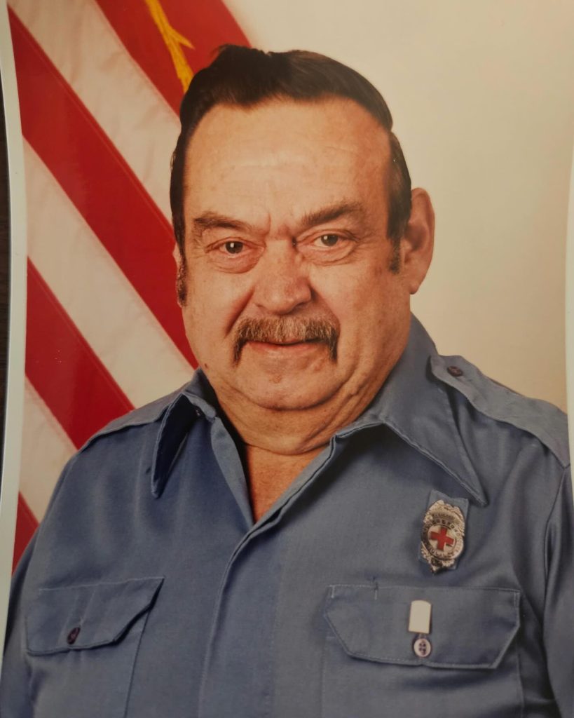 An older man with dark hair and a mustache wears a blue uniform shirt with a badge. He stands in front of an American flag, looking at the camera and smiling slightly.