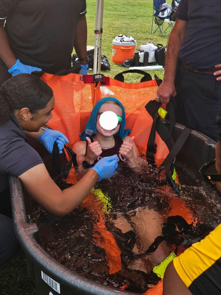 A person sits in a large tub filled with water and ice, giving two thumbs up, surrounded by medical staff wearing gloves. The person’s face is obscured, and outdoor supplies are visible in the background.